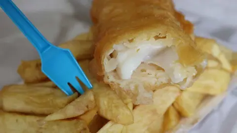 Reuters Close up of a classic portion of chip shop fish and chips. The fish is golden batter on the outside and white and flakey on the inside. A blue plastic fork is stuck in the chips