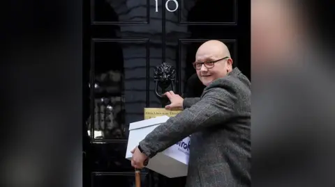 Stroke Association/PA Wire Campaigner Phil Woodford who is bald and wearing glasses and a tweed blazer knocks on the door of No 10 Downing Street with a box of signatures. He is smiling.