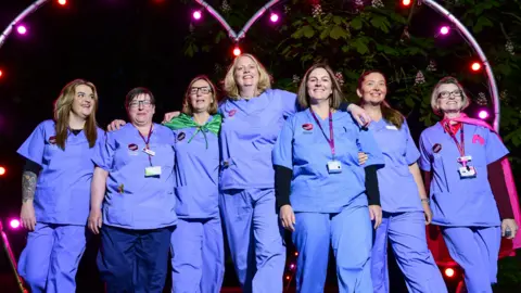 Nick Butcher Photography/St Elizabeth Hospice A group of hospice staff wearing matching blue medical scrubs stand in a line in front of a lit-up heart. They smile at the camera. 