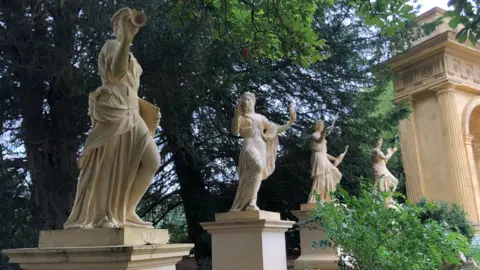 Martin Heath/BBC A row of pale stone statues stands on plinths in a landscaped garden beside a classical building with tall columns. The nearest statue holds a scroll and raises one arm, while the others hold objects including a mask and a long staff. Mature trees form a dense backdrop, with shrubs and gravel paths surrounding the sculptures. Sunlight filters through the leaves, illuminating the statues and the building's ornate façade.