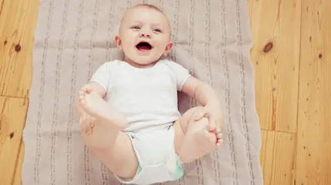 Getty Images A laughing baby in a white baby grow with a nappy underneath lies on a mat