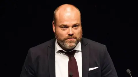 Getty Images Anders Holch Povlsen is a bald man with a brown beard. He is wearing a dark jacket, white short and brown tie