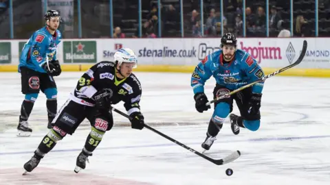 Getty Images Two blue uniformed ice hockey players skate towards a black uniformed player. A small crowd can be seen in the background behind a glass divider.  