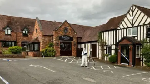 BBC The Pear Tree Inn. A red brick building with an officer is forensic PPE and police tape sealing off a black door into the building.