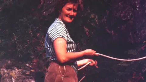 Rob Matheson Rob Matheson's mum Rena stands at the foot of a crag body holding on to a rope which she is using for belaying. She is wearing brown trousers and a black and white stripy shirt. She has brown curly hair and is half smiling. 
