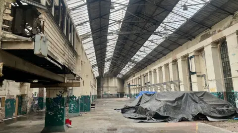 A large foundry with cream walls and pillars. Large equipment is covered by a grey tarpaulin. There is a burnt out office above the factory floor. There are three runs of clear panels to the roof allowing the light through. The walls are cracked and there are scribblings of graffiti.