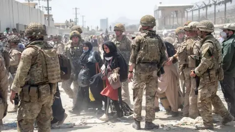 Getty Images British armed forces work with the US military to evacuate eligible civilians and their families out of Afghanistan on August 21, 2021. Soldiers on dusty road between wire fences topped with razor wire escorting two women dressed in black