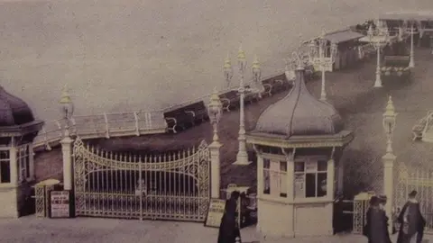 Openwide Coastal Ltd A black and white image of the old Cromer Pier. It has a gate and two small buildings either side of it at the entrance. On the pier are benches and lamp posts.