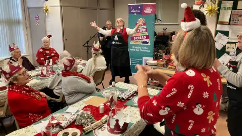 BBC/Piers Hopkirk People wearing Christmas party hats sit at dining tables as a woman wearing a kitchen apron stands at the front with her arms raised. Others watch on from the side.