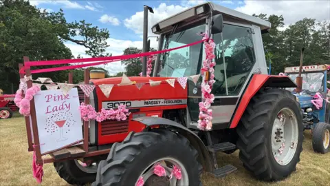 Richard Knights/BBC A small red tractor parked in a field. It has been decorated in pink bunting and flowers. There is a white sign attached with the picture of a pink cocktail on it and the words: Pink Lady.