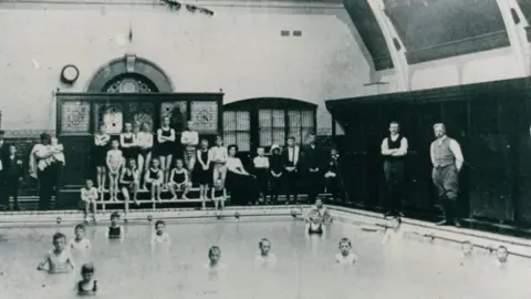 LDRS An early black and white picture of the inside of the baths. A number of children in swimming costumes are pictures sitting on benches next to the pool, with many also in the water. 