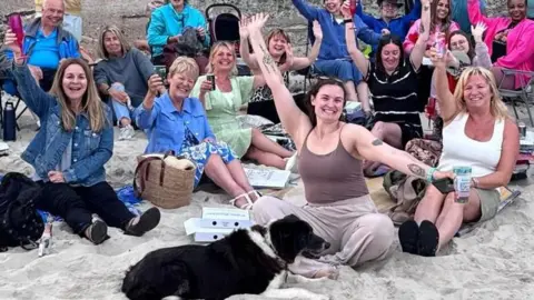 Large group of people seated and standing on a sandy beach in front of a concrete wall, raising their arms toward the camera. A dog lies on the sand in the foreground, with picnic items and bags scattered around.