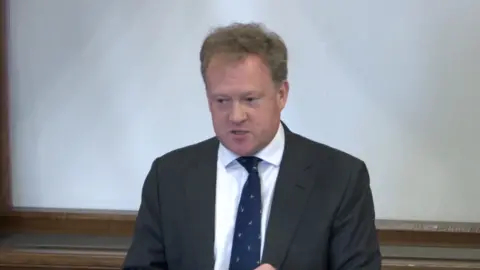 UK PARLIAMENT Greg Stafford, the Farnham and Bordon MP, talking in a debate at Westminster Hall in Parliament. He is wearing a suit and blue tie.