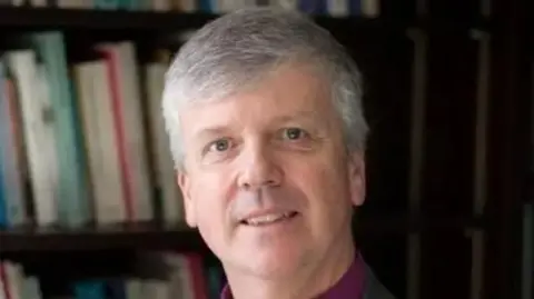 Andrew Watson, Bishop of Guildford, wearing dog collar and grey jacket in front of a bookshelf 