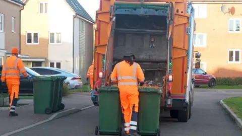 A red refuse collection lorry with two green bins at the back - ready to be unloaded into it. Crew members wearing orange high-visibility uniform can be seen dragging the bins.