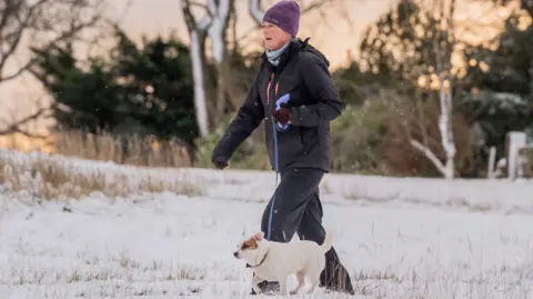 A woman wearing a purple hat, blue scarf and black outdoor gear, walks a small white and tan terrier-like dog through a snowy field, as the sun sets in the background 