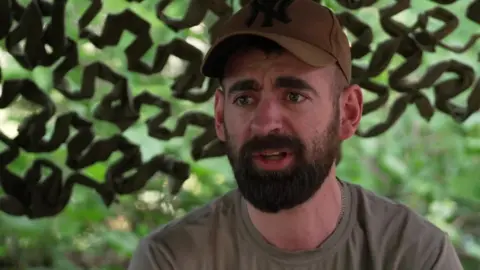 A man in an olive T-shirt and baseball cap sits in front of camouflage netting