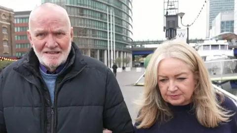 Tom Shaw and Kirstie Moore photographed while speaking to TV cameras. They are outside on a grey day. 