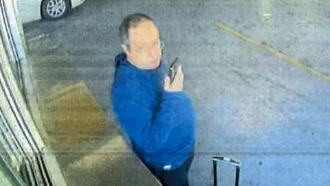 A person holding a phone and wearing a blue jumper stands next to a wheeled suitcase in an underground car park with cars in the background.