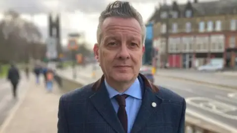 Nick Kemp, with short gray hair, stands on a main street. He wears a blue suit and looks expressionlessly into the camera.