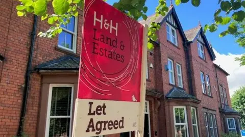 A letting sign featuring "H&H" in white letters, "Land and Estates" in black, both against a maroon background, with "Let Agreed" below on a white background. It is in front of a large brick built apartment block with bay windows on the ground floor.
