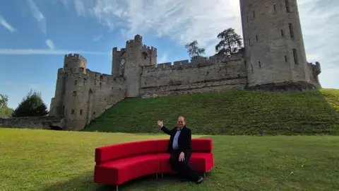 Reporter Ben Sidwell sitting on the red sofa with the backdrop of Warwick Castle behind him