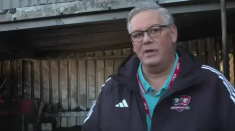 A man, Exeter City manager Clive Harrison in black coat, wearing glasses