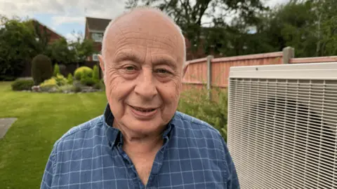 Dr David Toke standing in a garden. There is a heat pump next to him. He is balding with white hair and he is smiling. He is wearing a blue checked shirt which is open at the neck.