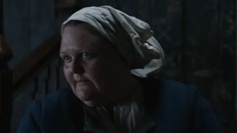 Dark shot of Laura Guest in period costume staring in Hamnet as a midwife. 