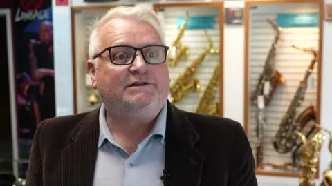 Walker stands in his music shop, speaking to a reporter. He has silver hair and has glasses. He wears a shirt and suit.