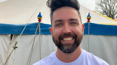 A man in a white t-shirt smiles in front of a tent. He has dark hair, blue eyes and a short beard. In the distance, a tree can be seen over his left shoulder.