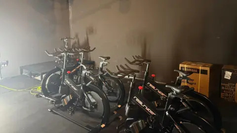 Image contains four black spin bikes clustered together, and in the background one more black spin bike is sitting on a raised platform for where the instructor will be. The floors and walls are painted black. There are two delivery boxes behind the bikes.
