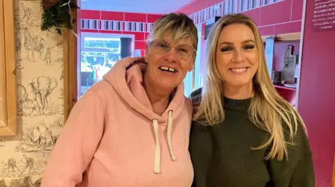 (left to right) Mags Fortune and Sharn Smith are standing indoors close together. Both are smiling at the camera. Mags is wearing a light pink hoodie, and Sharn is wearing a dark green jumper. They are standing in front of red walls on one side and a wall with wallpaper and animal illustrations, including elephants and zebras.