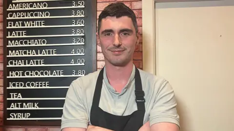 A man is standing next to a coffee menu that's attached to a wall in his café. His arms are folded and he's wearing an apron. There's a coffee machine in the corner. 