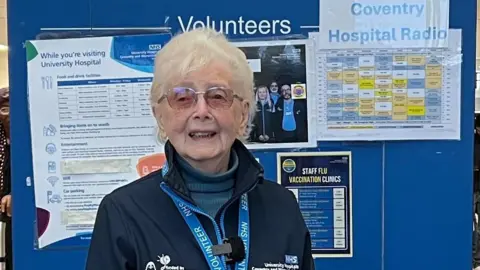 Betty Miller standing at the information desk in the lobby of Coventry's University Hospital. She has white hair and is wearing her blue volunteer's uniform with a volunteer lanyard. She is standing in front of a blue sign marked "Information Desk".