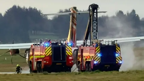 Two airport fire trucks with flashing blue lights positioned behind a turboprop aircraft on a grassy area, with smoke or steam rising near the plane and a firefighter working on the ground