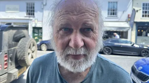 Ben Parker/BBC A man looking at the camera, he has a beard and a blue top on, cars are parked in the background on the street.