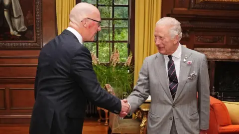 PA Media John Swinney wearing a dark coloured suit shakes the hand of King Charles wearing a light grey suit and black striped tie.