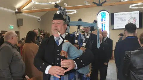 A piper is playing his instrument. He's wearing a kilt. It's a blue bagpipe and with four pipes. OPne is in his mouth. He's leaving a church. People carrying a coffin are behind him. A congregation is standing. 
