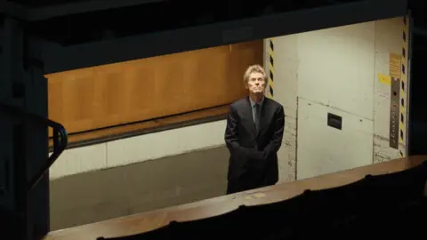 Manchester Film Festival handout Actor Willem Dafoe in a scene from The Souffleur film. He is wearing a suit and is standing in a dugout type room. 