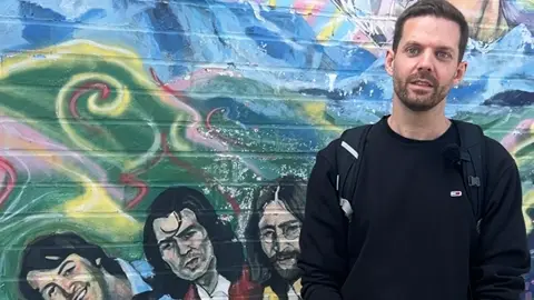 Ian van de Waal stands in front of a colourful mural showing three of The Beatles. Ian has short dark hair and a short beard. He wears a black jumper and carries a rucksack.