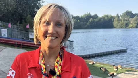 Sue Merby, who is wearing a red T-shirt and has two medals around her neck, is smiling at the camera in front of a reservoir. 