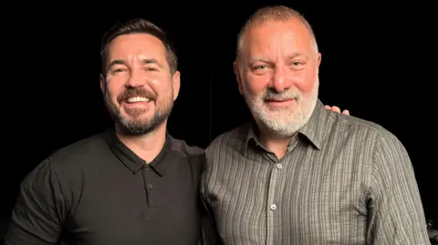 Martin Compston, wearing a black shirt, and Jed Mercurio - who has a white beard and a grey and black patterned shirt - smile into the camera. Compston has his arm around Mercurio.