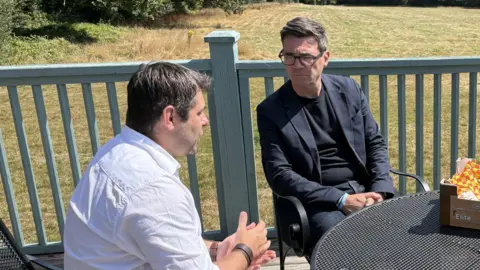 Andy Burnham wearing a dark top and jacket sitting next to Leigh Frost on his recent visit to Cornwall 