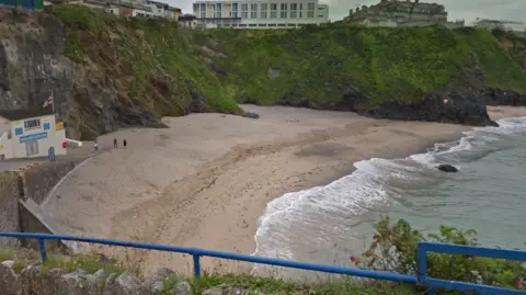 A heightened view looking down on to Great Western beach. It is an overcast day, there are cliffs at the back of the beach which are covered in grass and moss. The tide is halfway out and there are two small figures walking on to the beach. Above the cliffs are a series of buildings. 