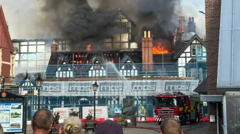 Richard Morris An old white building with black timber frames surrounded by scaffolding with flames and smoke coming from the roof