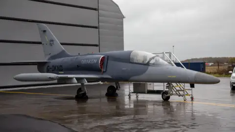 Tees Valley Combined Authority A Draken fighter jet parked outside a hangar at Teesside Airport. The grey aircraft has a long nose and a small cockpit. 