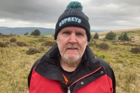Geraint Evans, who is campaigning against wind and solar farms in the Dulais valley, stands on moorland wearing a red and black raincoat and wearing a black woollen hat which says 'Ospreys' on it. It is a head and shoulders shot.