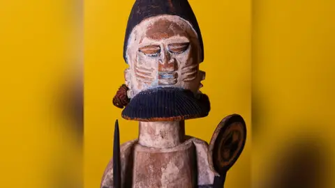 Manchester Museum handout Carved wooden figure on show at an exhibition with a yellow background.