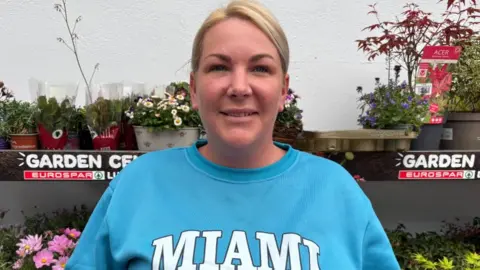 A woman with blonde tied back hair smiles into the camera. She has a nose ring and is wearing a light blue sweatshirt that says 'Miami' in capital letters. She is standing in front of a stall of flowers in plastic pots.